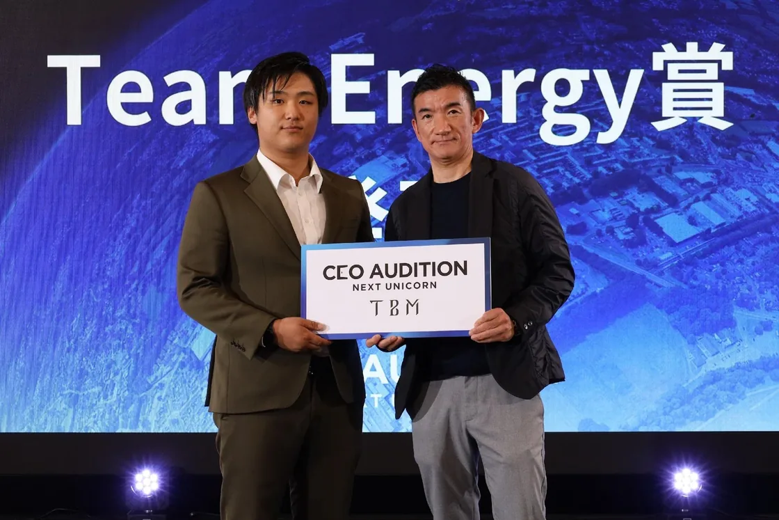 Two men on a stage holding a board that reads CEO Audition NEXT Unicorn, in front of a screen announcing the prize.